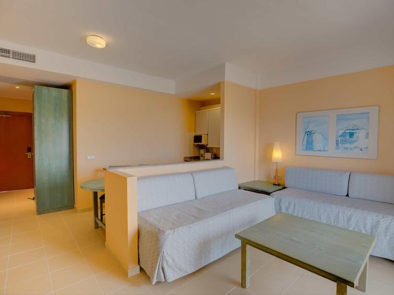 Camera d'hotel luminosa con due divani, tavolo in vetro e arte murale minimalista.