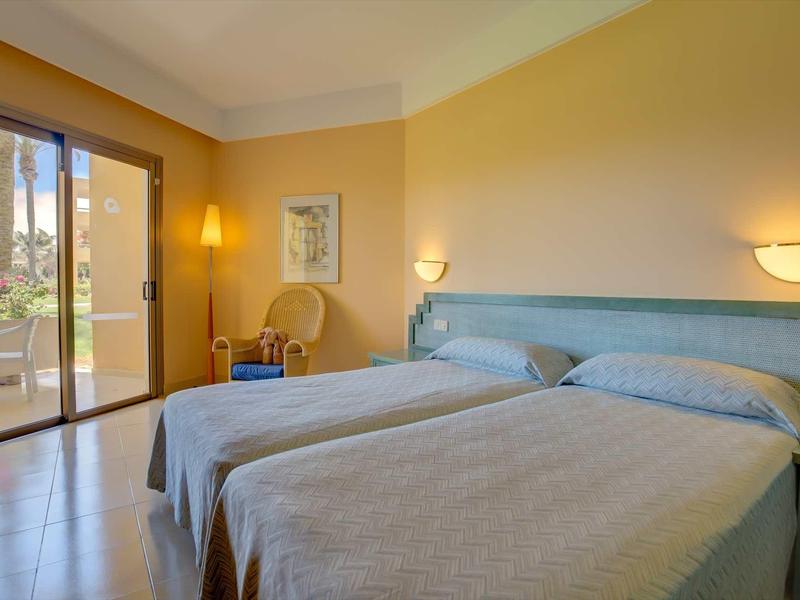 Camera d'hotel luminosa con due letti singoli e accesso a una terrazza con vista sulle palme.