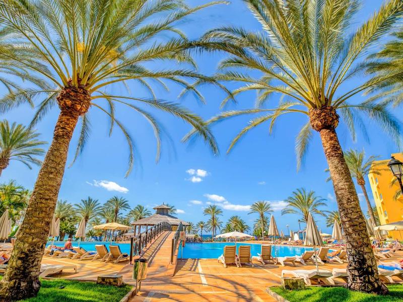 Area piscina con palme e lettini sotto un cielo azzurro in un resort.
