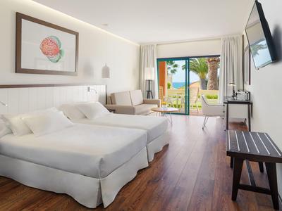Bright hotel room with two beds, seating area, and view of tropical garden.