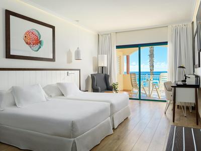Bright hotel room with two beds, balcony, and sea view in natural light.