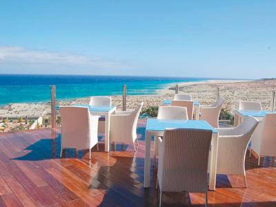 Terrace with white chairs and tables overlooking the sea and coastline under clear sky.