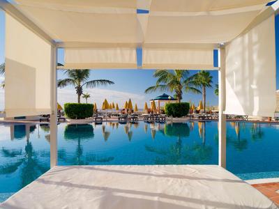 Vista desde una cabaña sombreada hacia una piscina con palmeras y sombrillas.