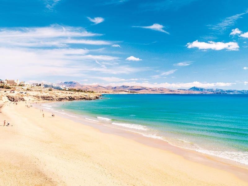 Spiaggia sabbiosa soleggiata con acqua turchese chiara e cielo azzurro con nuvole.
