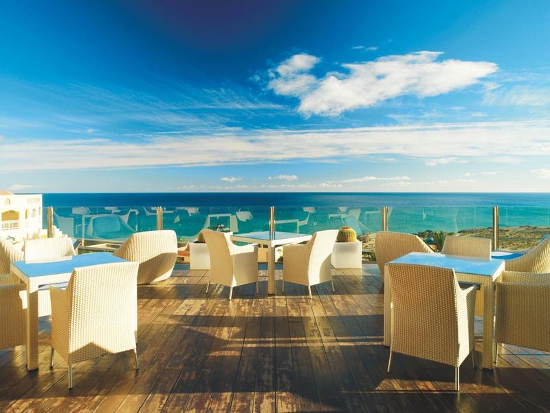 Area pranzo all'aperto con sedie bianche e tavoli con vista sul mare e cielo azzurro.