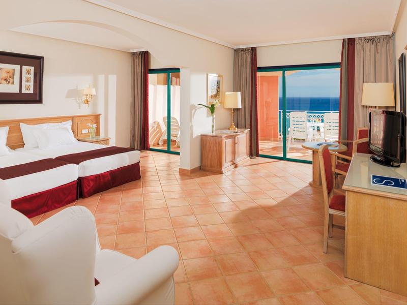 Camera d'hotel luminosa con due letti, scrivania, poltrona e balcone con vista sul mare.