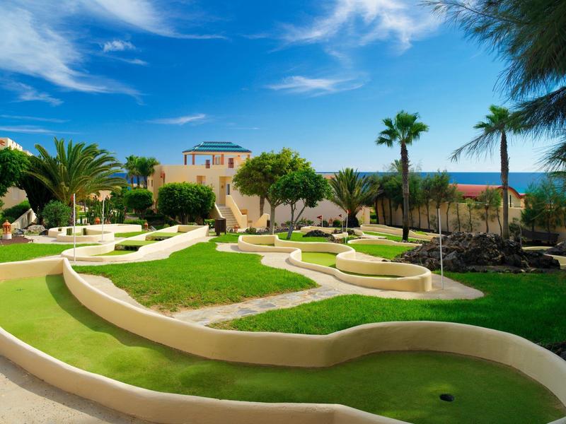 Percorso da mini golf all'aperto con prati verdi, palme e edificio vicino al mare sotto un cielo blu.