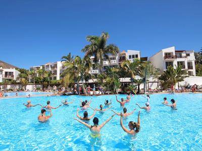 Menschen machen Wassergymnastik im blauen Pool vor weißen Ferienhäusern und Palmen bei sonnigem Wetter.