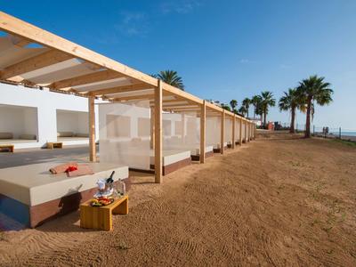 Moderner Strandpavillon mit Holzdach, weißen Sitzbereichen und Palmen unter klarem blauem Himmel.