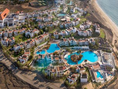 Ansicht einer großen Hotelanlage mit blauen Pools, grünen Gärten und angrenzendem Strand am Meer.