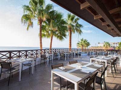 Terrassenrestaurant mit weißen Tischen, schwarzen Stühlen und Palmen am Meer bei Tageslicht.