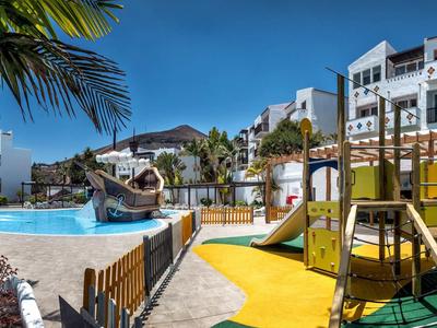 Bunter Kinderspielplatz neben rundem Pool mit Rutsche, Palmen und weißem Wohngebäude im Hintergrund.