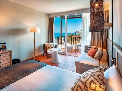 Komfortables Hotelzimmer mit Meerblick, hellem Holz, Sofa, Bett und Balkon mit Korbstühlen.