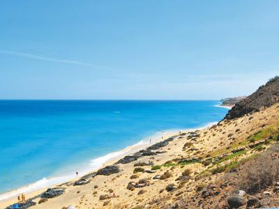 Plage avec sable doré, rochers et mer bleue claire sous un ciel bleu.
