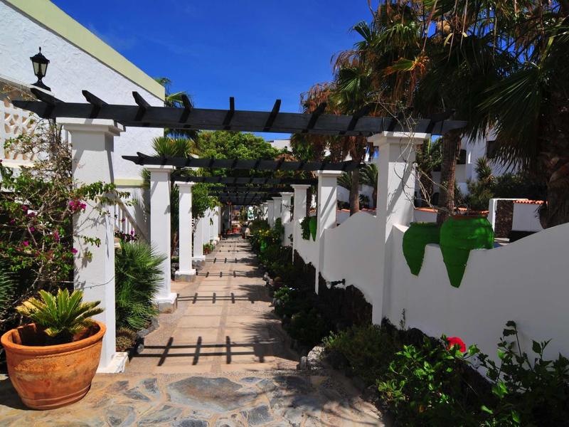 Chemin ensoleillé avec pergolas et murs blancs, entouré de plantes dans un jardin d'hôtel.