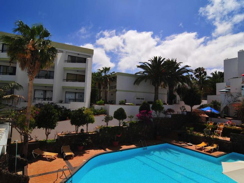 Hôtel avec piscine extérieure, entouré de palmiers et de plusieurs bâtiments blancs sous un ciel bleu.