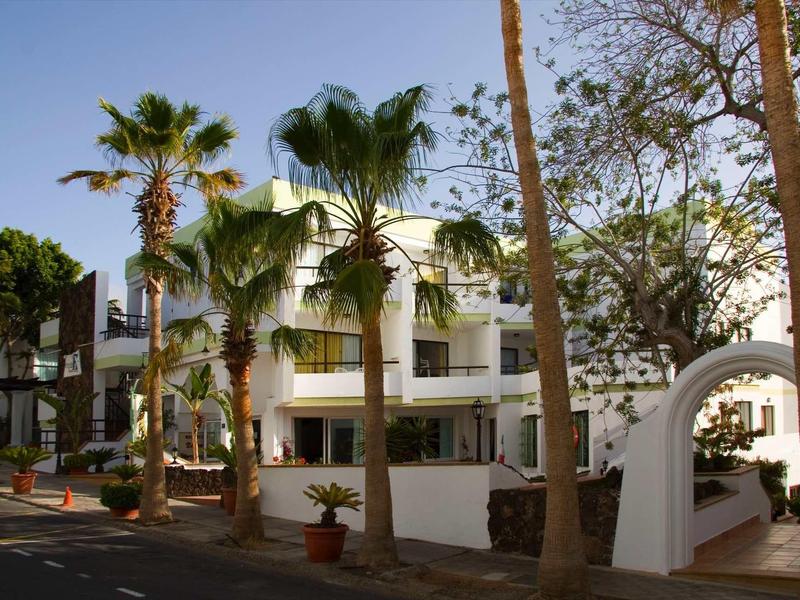 Bâtiment blanc avec balcon entouré de palmiers et d'arbres sous un ciel clair.