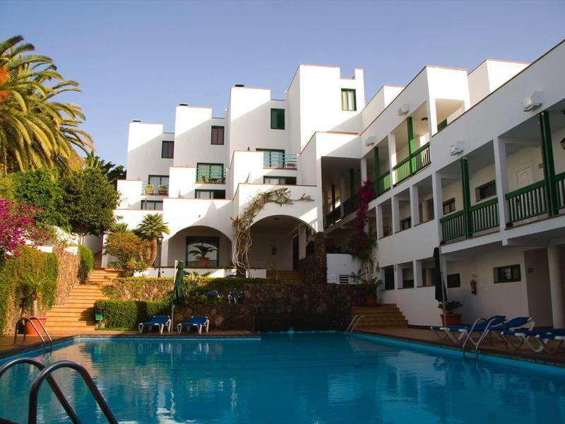 Complexe hôtelier moderne avec piscine extérieure, chaises longues et palmiers sous un ciel bleu.