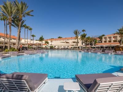 Großer Hotelpool mit Liegestühlen und Palmen unter klarem, blauem Himmel.