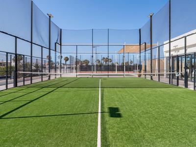 Court de padel moderne avec gazon synthétique vert et filets, entouré de bâtiments et ciel bleu.
