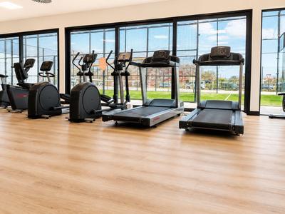 Heller Fitnessraum mit Cardiogeräten vor bodentiefen Fenstern und Blick auf Außenbereich.