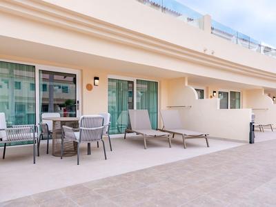 Terrasse mit Tisch, Stühlen und Liegestühlen vor modernen Hotelzimmern an sonnigem Tag.