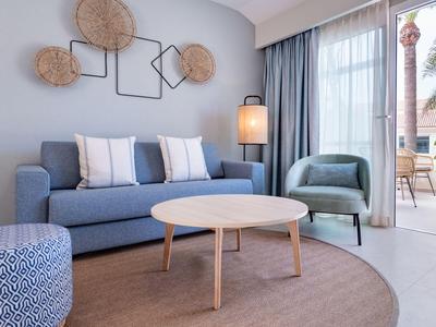 Hell eingerichtetes Wohnzimmer mit blauem Sofa, Sessel und Zugang zur Terrasse mit Palmenblick.