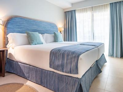 Helles Hotelzimmer mit großem Bett, blauer Bettwäsche und Vorhängen neben Fenster.