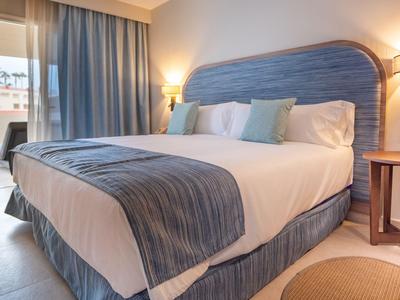 Gemütliches Doppelbett mit blauem Kopfteil und Vorhängen in einem Hotelzimmer mit Balkon.