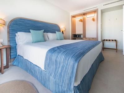 Modernes Hotelzimmer mit weißem Bett und blauer Bettdecke, Beistelltischen und Garderobe.