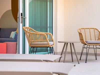 Zwei Rattanstühle und ein kleiner Beistelltisch auf einer hellen Terrasse mit Blick auf ein geöffnetes Schiebetürfenster.