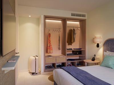 Modern hotel room closet with open wardrobe, clothing, suitcase, bedside lamp, and bed with pillows.