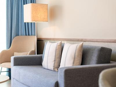 Modernes Hotelzimmer mit grauem Sofa, zwei Kissen, Lampe und beigem Sessel neben blauen Vorhängen.