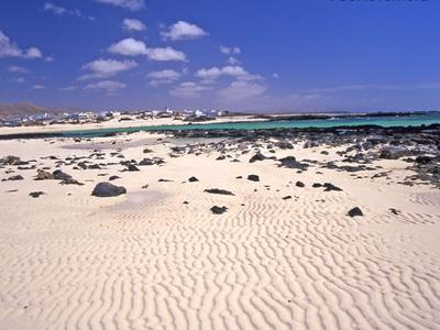 Grande étendue de sable avec des rochers noirs près de la mer bleue sous un ciel clair à Fuerteventura.