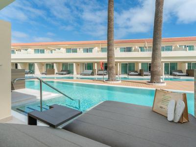 Espace piscine moderne avec chaises longues et palmiers devant des chambres d'hôtel sous un ciel bleu.