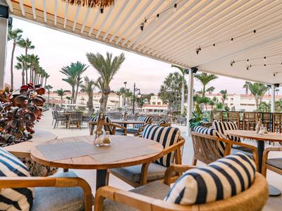 Terrasse couverte d'un hôtel avec des tables en bois, des chaises rembourrées et vue sur des palmiers.