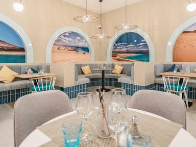 Elegante Hotelbar mit runden Tischen, gemütlichen grauen Sesseln und wandfüllenden Strandmalereien.