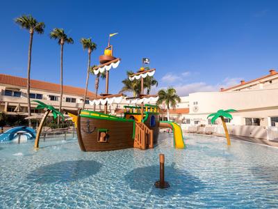 Piscine pour enfants avec aire de jeux en forme de bateau pirate, toboggans aquatiques et palmiers dans un complexe hôtelier.