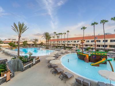 Grand piscine d'hôtel avec palmiers, chaises longues et aire de jeux aquatique sous un ciel bleu.