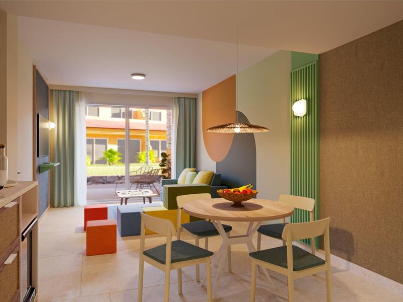 Bright modern hotel room with dining table, chairs, sofa, and balcony with outdoor seating.