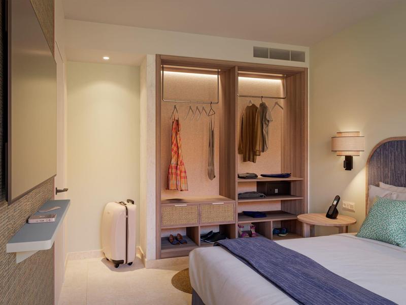 Modern hotel room closet with open wardrobe, clothing, suitcase, bedside lamp, and bed with pillows.