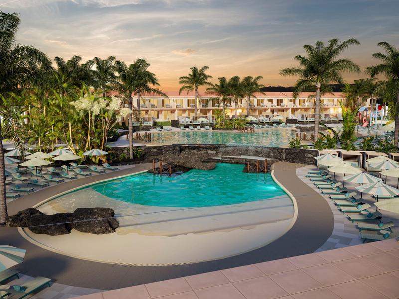 Resort pool area with palm trees, sun loungers, and a bridge during sunset.