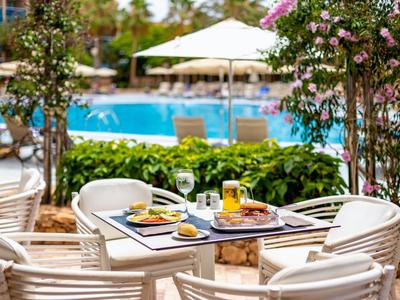 Esstisch mit vier Stühlen vor einem Pool, umgeben von grünen Pflanzen und Sonnenschirm.