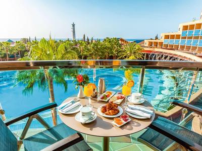 Table sur balcon d'hôtel avec petit déjeuner donnant sur piscine et palmiers sous ciel dégagé.