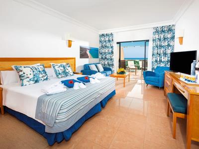 Chambre d'hôtel lumineuse avec deux lits, accents bleus et vue sur la mer depuis le balcon.