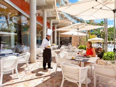 Un serveur sert des boissons à une femme sur une terrasse ensoleillée avec des chaises et des tables blanches.