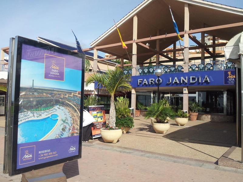 Entrée d'hôtel avec enseigne 'Faro Jandia' et panneau publicitaire avec photo de plage