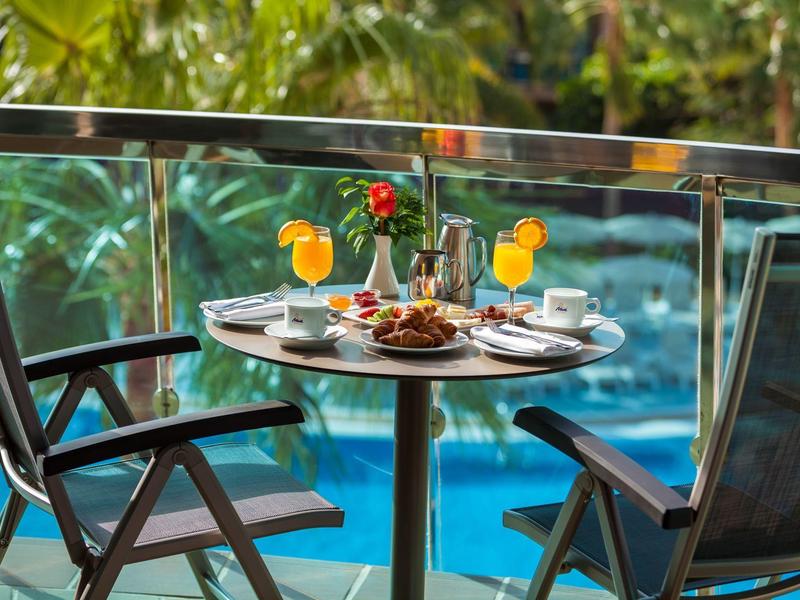 Table de balcon avec nourriture, boissons et deux chaises avec vue sur une piscine et des plantes tropicales.