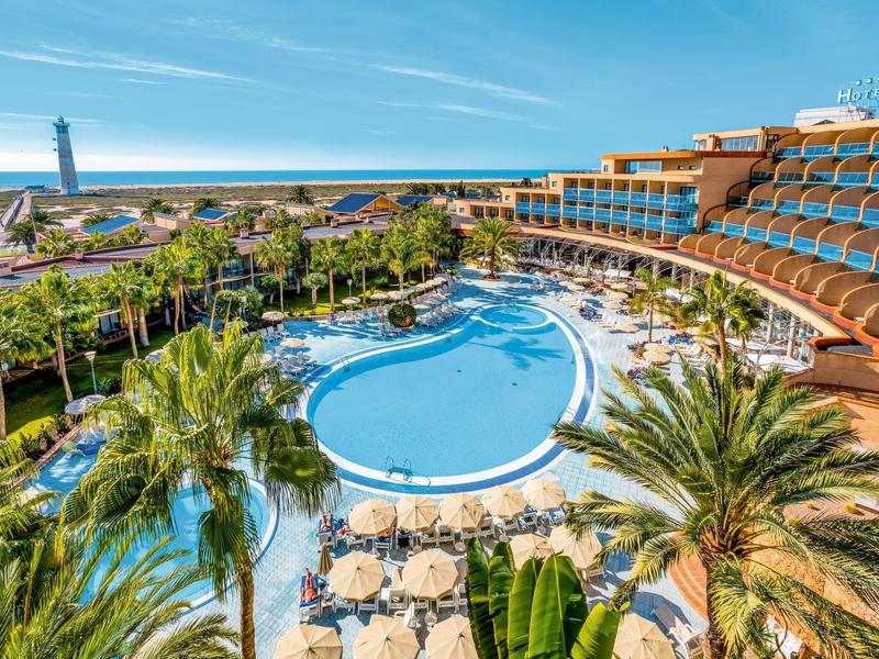 Vue d'un hôtel avec une piscine circulaire, des parasols et des palmiers sous un ciel bleu.