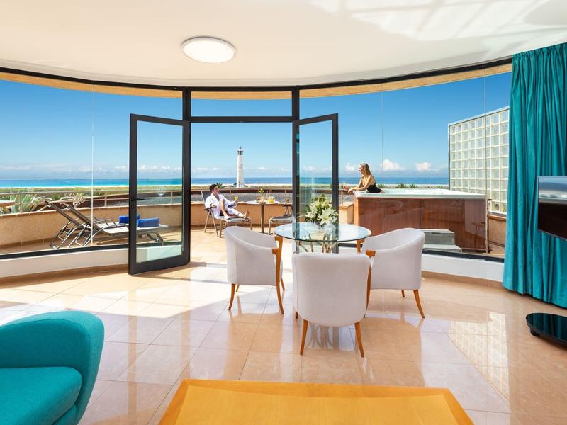 Chambre d'hôtel lumineuse avec balcon, fenêtres panoramiques et vue sur la mer.